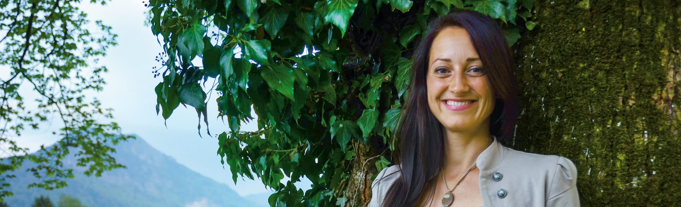 Nicole Künzl lächelt neben einem efeubewachsenen Baumstamm, dahinter Berge