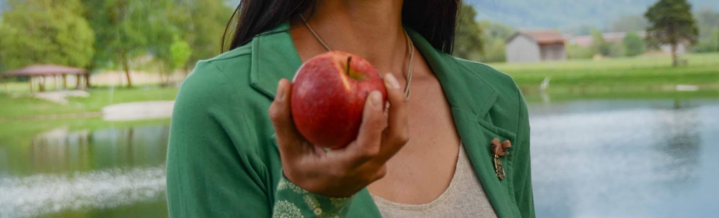 Hand in grüner Jacke, die einen roten Apfel vor einem See hält